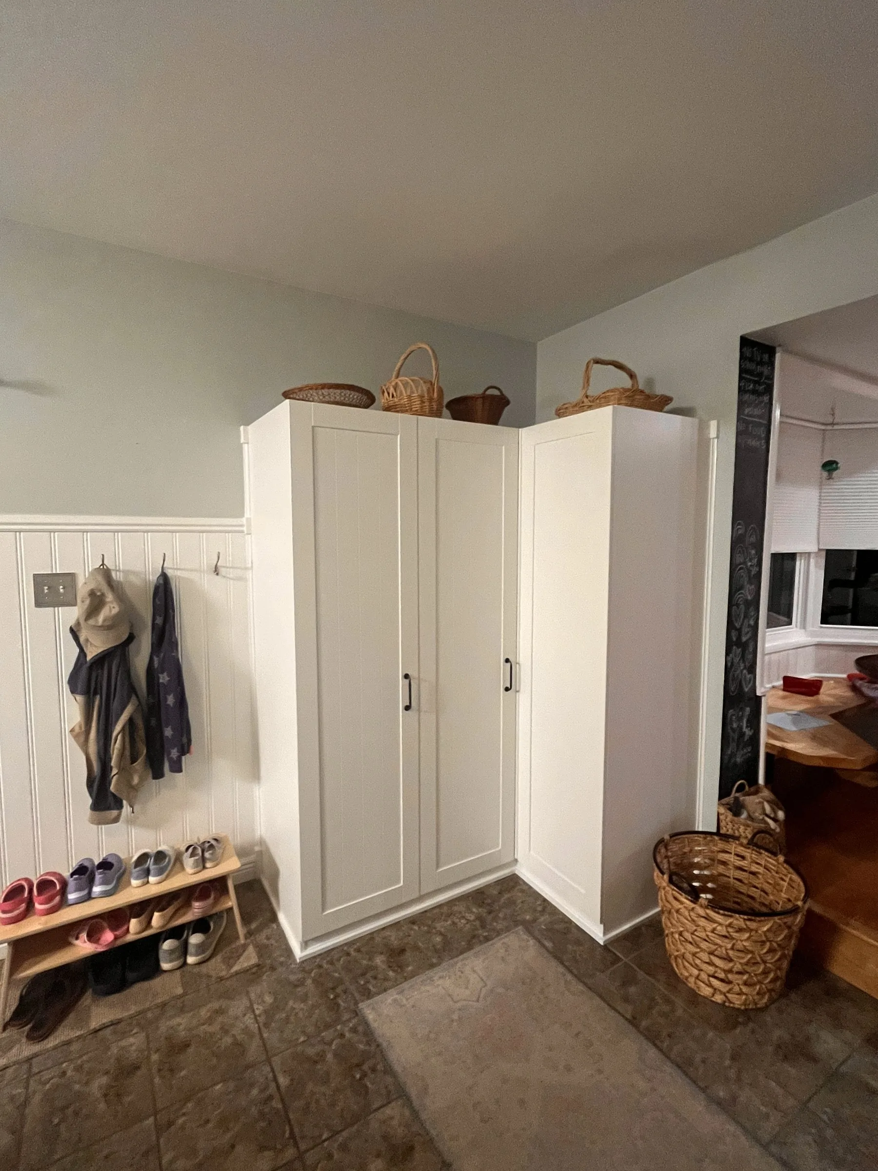 Two tall white corner cabinets in a mudroom, baskets on top, beadboard wainscot and coat hooks to the left