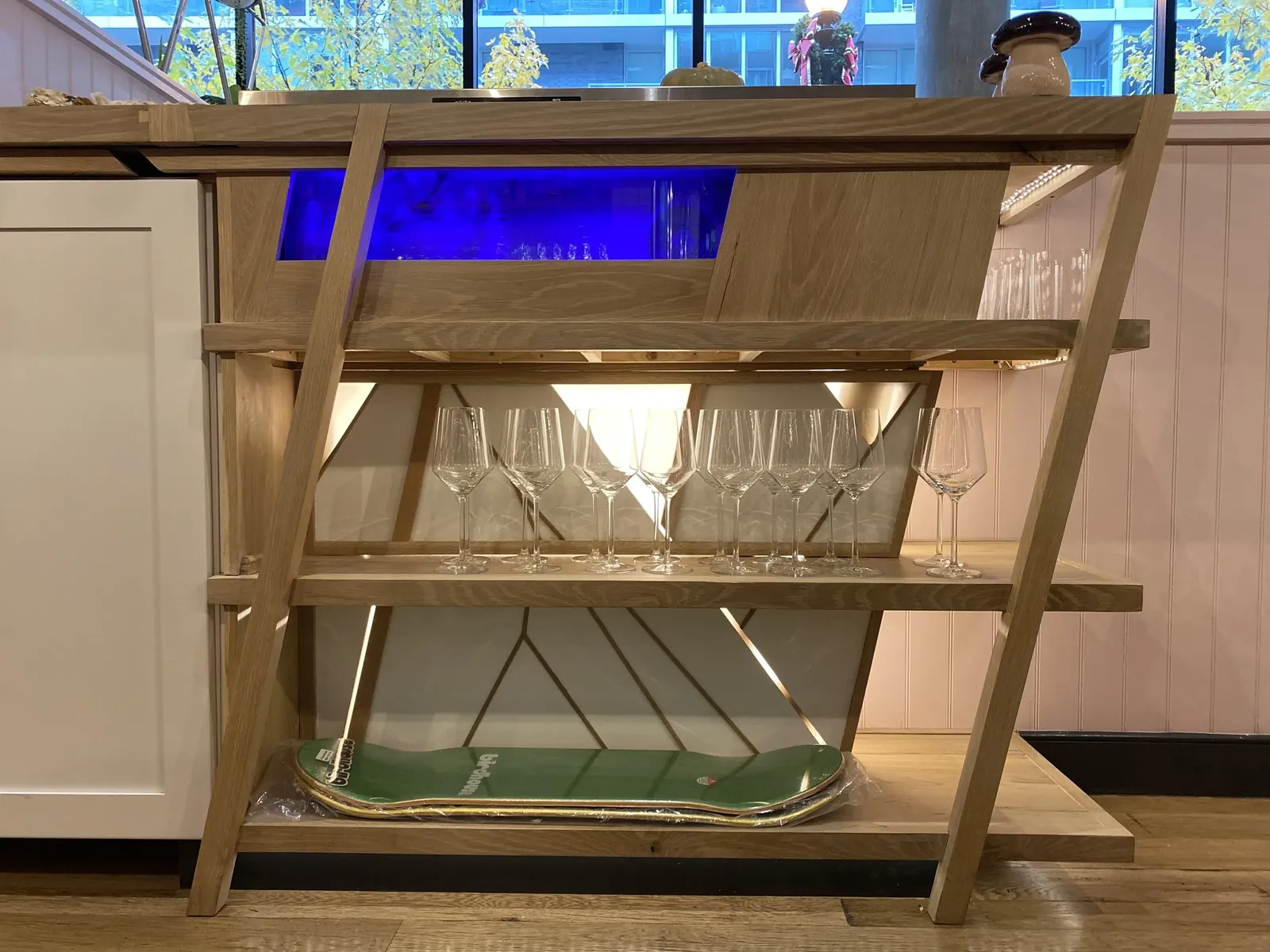 Angled light-wood shelf with wine glasses and a skate deck on the bottom